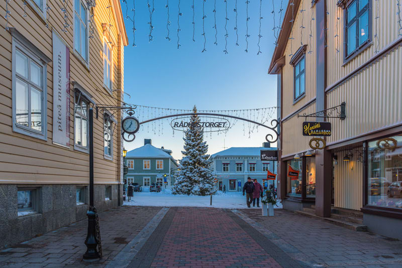 Bilden visar Rådhustorget i Piteå under vintertid. Till vänster i förgrunden syns en gul träbyggnad med banderollen "piteå museum" vertikalt längs fasaden. En dekorativ svart lyktstolpe med en klassisk klocka är monterad på byggnaden. I mitten av bil