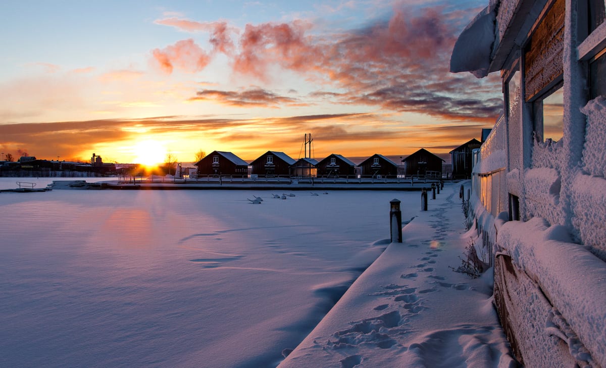 Bilden visar ett vinterlandskap vid solnedgången. En frusen sjö eller ett vattendrag täckt av snö sträcker sig över förgrunden och mellangrunden. I bakgrunden syns en rad mörka hus, troligen sjöbodar eller småstugor, med snö på taken. Till höger i bi