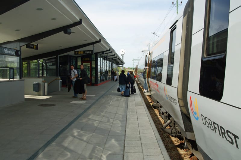 Bilden visar en modern tågperrong utomhus under dagtid. Till vänster finns en byggnad med delvis glastak och skyltar, varav en indikerar "Falkenberg" och en annan "Utgång". På perrongen står flera personer med bagage, varav några väntar och andra ver