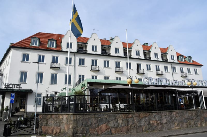 En bild visar Grand Hotel i Lundby beläget utomhus under dagtid. Hotellet är en vit byggnad med ett rött tak. Framför bottenvåningen finns en uteplats med svarta bord och stolar. Ovanför entrén syns hotellets namn: 'GRAND HOTEL'. På fasaden till väns