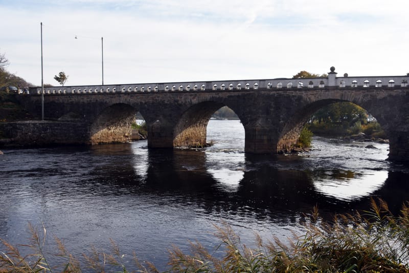 Bilden visar en stenbro med flera valv som sträcker sig över en vattendrag under dagtid. Bron är byggd av grå sten med en vit räckeskonstruktion ovanpå som har dekorativa öppningar. Vattnet under bron reflekterar delar av brons struktur. På brons bor