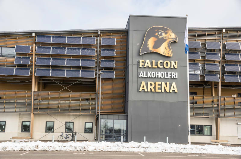 Bilden visar utsidan av en byggnad kallad "FALCON ALKOHOLFRI ARENA". Byggnaden har en grå fasad på den högra sidan, där arenans namn och en logotyp föreställande ett falkhuvud är placerade. Den vänstra delen av byggnaden har en fasad av horisontella