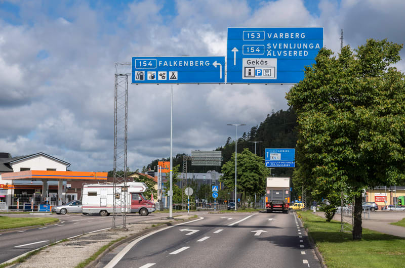 Bilden visar ett landskap med en väg i förgrunden som leder in i bilden. Ovanför vägen hänger två blåa vägskyltar på en metallkonstruktion. Skyltarna indikerar riktningar till Falkenberg (vänster) samt Varberg, Svenljunga, Älvsered och Gekås (rakt fr