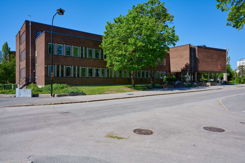 Bilden visar en större byggnad i tegel med många fönster längs fasaden, placerad bredvid, och delvis bakom, ett större lummigt träd med gröna löv. Byggnaden är kommunhuset i Tierp