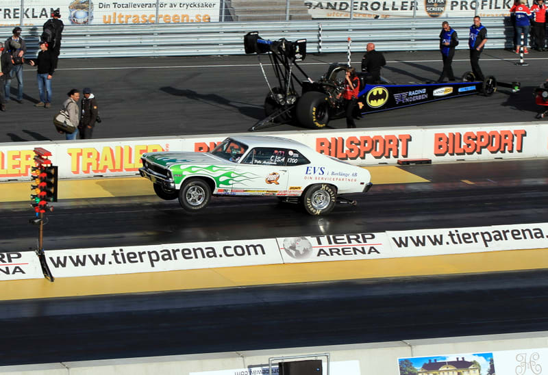 En bild tagen utomhus under dagtid på en racingbana. I förgrunden till vänster syns en startlampa för dragracing med röda, gula och gröna lampor. En vit bil med gröna lågor målade på sidorna accelererar från startlinjen med framhjulen lyfta. Bilen ha