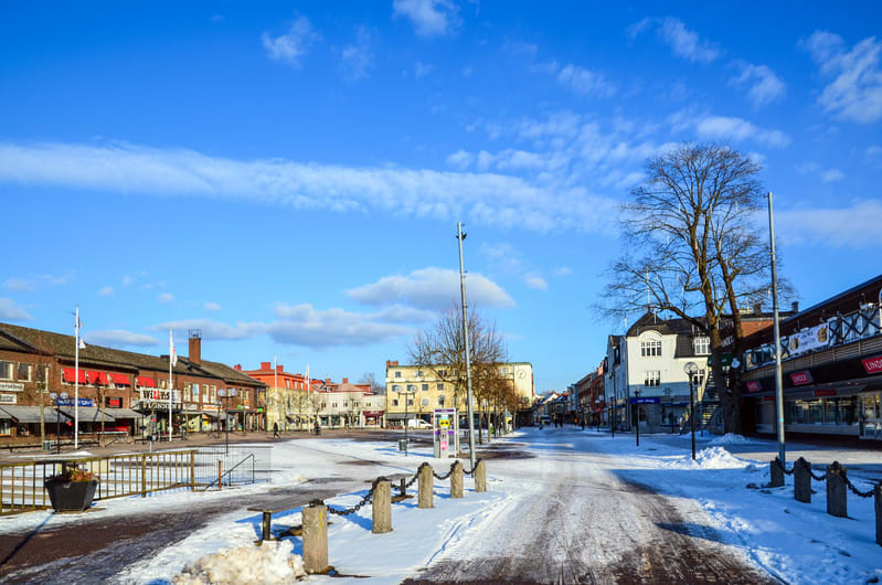En bild tagen från gatumarkhöjd en solig vinterdag som visar ett torg eller en öppen plats i en stad. Marken är täckt av snö och det syns spår av trafik på en del av marken. Runt torget finns byggnader i varierande höjd och färg, med butiksfasader oc