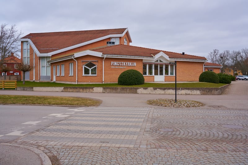 En bild tagen utomhus visar Pingstkyrkan Rydebäck under en mulen dag. Byggnaden är till största delen byggd i rött tegel med ett tegeltak i liknande nyans. Vita detaljer syns runt fönster och takfot. På byggnadens framsida finns en skylt med texten '