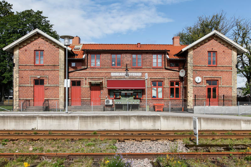 En fasadbild av en äldre tegelbyggnad, förmodligen en järnvägsstation, under en sommardag. Byggnaden är huvudsakligen konstruerad av rött tegel med ljusare tegeldetaljer och har ett tak med röda takpannor. Fönsterkarmar och dörrar är målade i en star