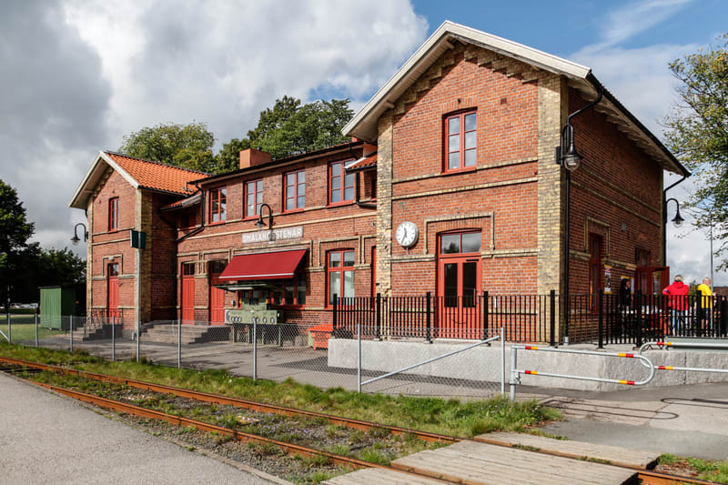 Bilden visar en tegelbyggnad med orangefärgade takpannor, vita takfotsbrädor och röda fönsterkarmar och dörrar. På byggnaden sitter en röd markis med text under, samt en skylt som delvis är skymd men där "SMÅLANDS STENAR" kan utläsas. En klocka är mo