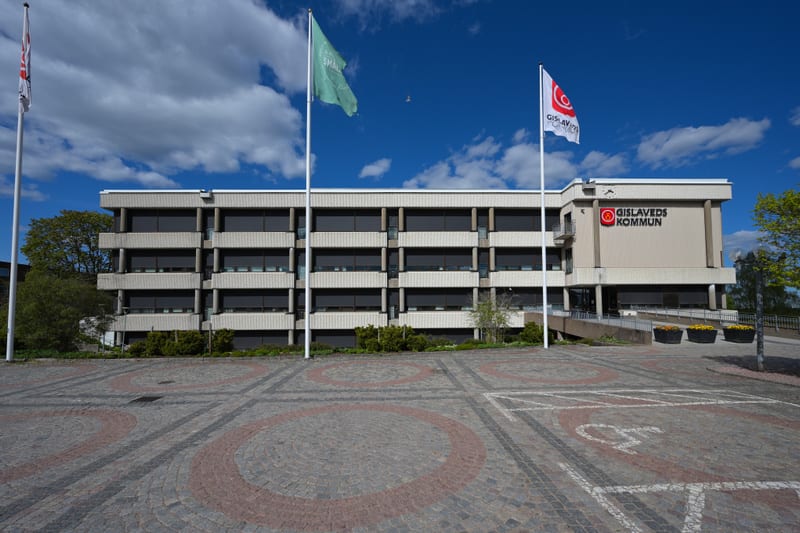 En fasadbild av Gislaveds kommunhus under dagtid. Byggnaden är beige och brun med svarta fönsterjalusier och flera våningar. Framför byggnaden finns ett stenlagt torg med cirkulära mönster i rött och grått. Två flaggstänger reser sig på torget, varav