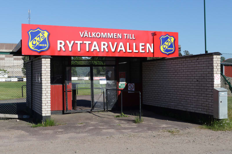 Bilden visar entrén till "Ryttarvallen" under en solig dag utomhus. Ovanför entrén finns en stor röd skylt med texten "VÄLKOMMEN TILL RYTTARVALLEN" i vitt. På sidorna av texten finns två gulblåa emblem med "GJS 1903" skrivet inuti. Entrén består av e