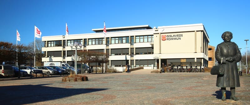 En stor, ljusbeige byggnad med mörka fönster och rullgardiner syns under en klarblå himmel. På byggnadens framsida står texten "GISLAVEDS KOMMUN" med en logotyp. Framför byggnaden finns en stor öppen yta med marksten i mönster. Till vänster om byggna