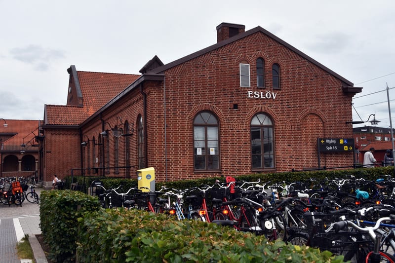 En bild som visar en tegelbyggnad, troligen en stationsbyggnad, med texten 'ESLÖV' på fasaden. Byggnaden har bågformade fönster och dörröppningar. Framför byggnaden finns ett stort antal parkerade cyklar, avskärmade av en grön buskhäck. En gul parker