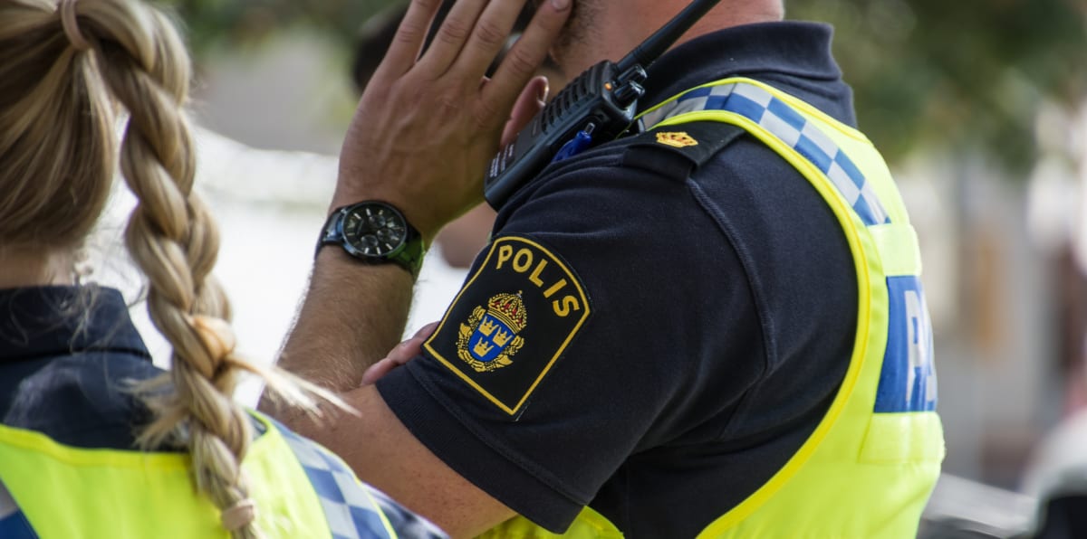 Bilden visar delar av två personer, troligen poliser, utomhus under dagtid. I förgrunden syns bakhuvudet på en person med blont, flätat hår och en gul reflexväst. Till höger om denna person, och något i bakgrunden, syns en annan person klädd i mörkbl
