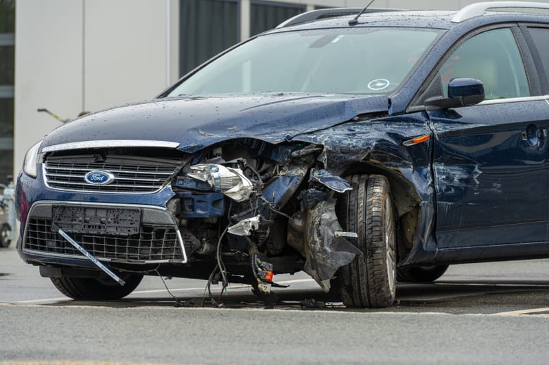Bilden visar fronten och högra sidan av en mörkblå personbil av märket Ford. Bilen har betydande skador på fronten, särskilt på vänster sida där stötfångare, grill och strålkastare är krossade och deformerade. Även motorhuven och vänster framskärm är