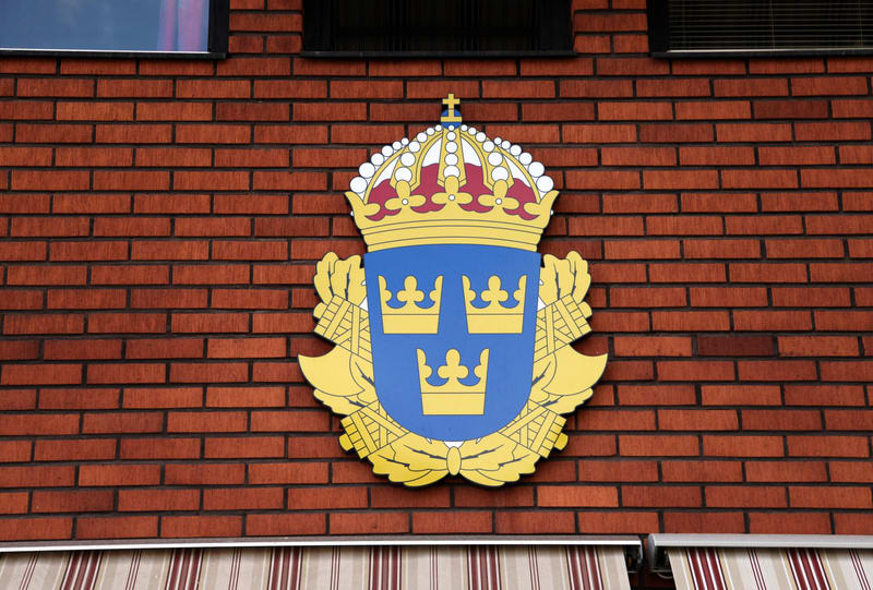 Bilden visar ett gult, blått och rött emblem med tre kronor som sitter monterat på en vägg av rött tegel. Emblemet, som liknar Sveriges lilla riksvapen, är centrerat i bilden. Ovanför emblemet syns ett fönster eller en mörk glasyta, och nedanför skym