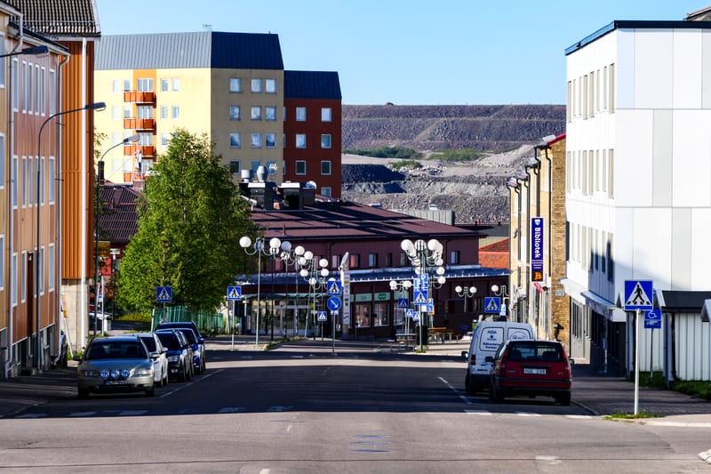 Bilden visar en gata med byggnader på båda sidor under dagtid med klarblå himmel. På vänster sida av gatan står flera bilar parkerade längs trottoaren. Längre bort på vänster sida syns en gul byggnad med balkonger och tak av mörk färg, samt en rödbru