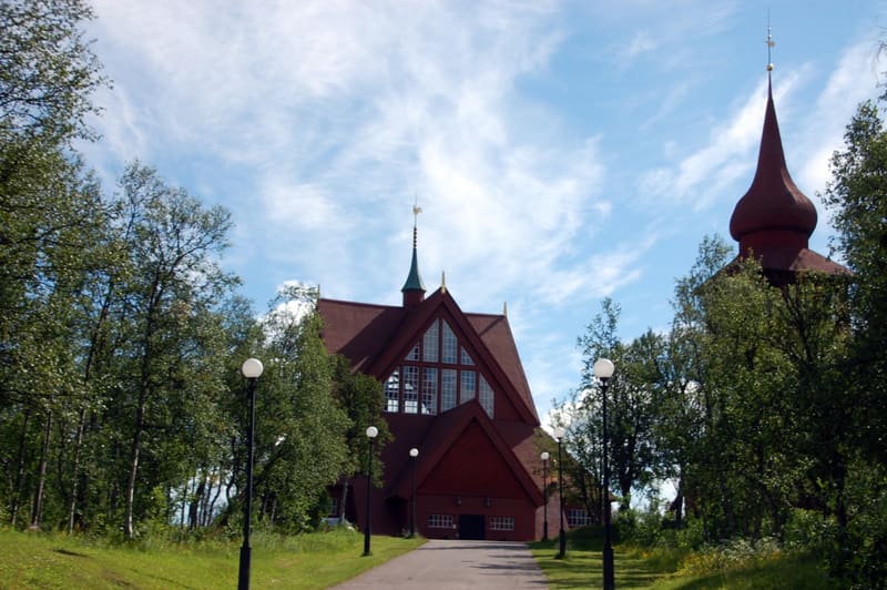 Bilden visar två byggnader med röda tak och fasader, omgivna av gröna träd och gräs. Den centrala byggnaden har en spetsig taknock med en vindflöjel och stora fönsterpartier i gaveln. En grusgång leder fram till denna byggnad, kantad av gatlyktor med
