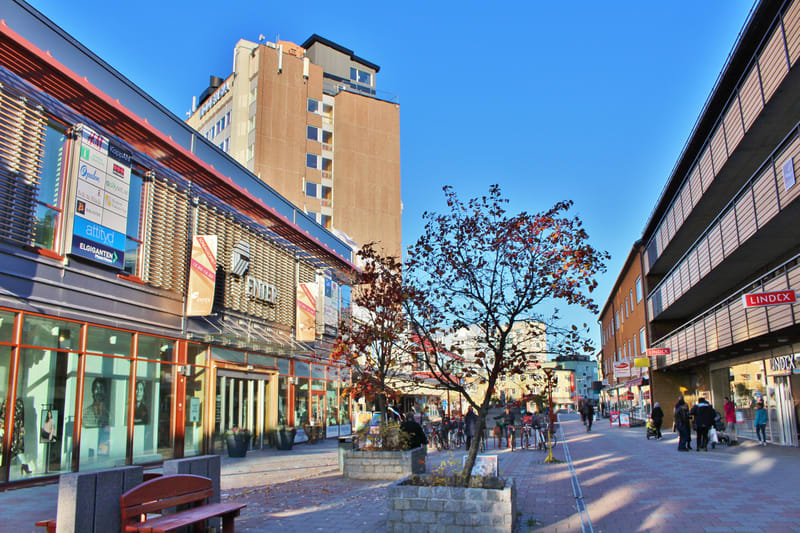 Bilden visar en gatumiljö med butiksfasader och människor. Längst till vänster syns delar av en träbänk. Till vänster i bilden finns en stor byggnad med skyltfönster och ingångar, samt skyltar för olika butiker som "Kopparhammaren", "attityd" och "EL