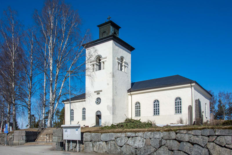Bilden visar en vit kyrka med ett fyrkantigt torn upptill, med ett svart tak och en korsformad topp. Kyrkan har flera fönster med rundade, bågformade överdelar. Framför kyrkan finns en stentrappa som leder uppåt och en stenmur som löper längs bildens