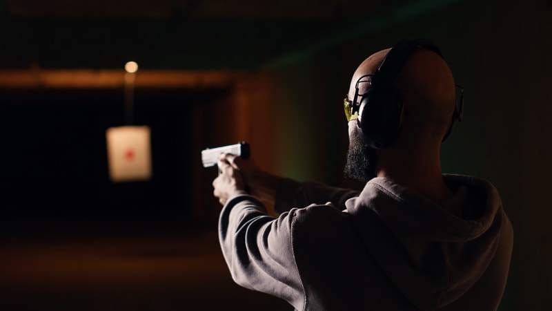 En person som bär hörselkåpor och gula skyddsglasögon håller i en handeldvapen och siktar mot en måltavla upphängd i bakgrunden. Personen är fotograferad bakifrån och har på sig en huvtröja. Bakgrunden är mörk med diffus belysning som skapar en gradi