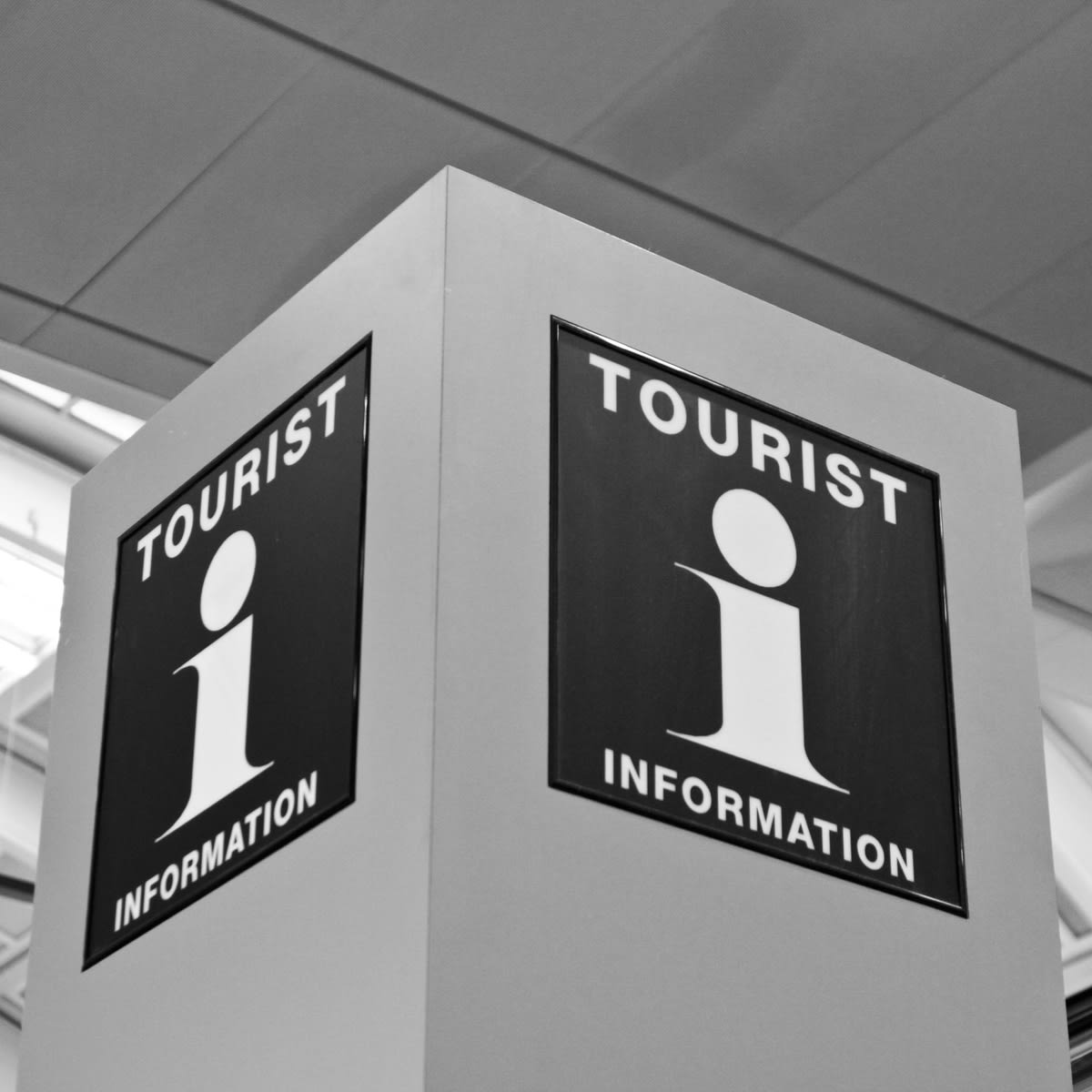 Bilden visar en skylt i svartvitt med information för turister. Skylten är rektangulär och har texten "TOURIST INFORMATION" i stora vita versala bokstäver, samt en stiliserad vit 'i' symbol på en svart bakgrund. Skylten är monterad på en ljusgrå pela