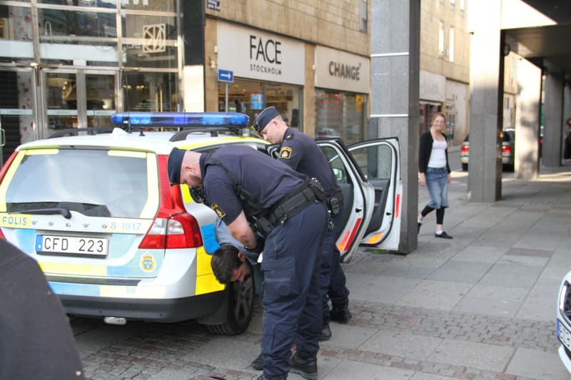 Bilden visar en gatascen där tre män och en kvinna är synliga. Två män i polisuniformer befinner sig vid bakre delen av en vit och gul/blå polisbil av märket Volvo. Den ena polismannen böjer sig fram mot en tredje man som verkar stå böjd med armarna