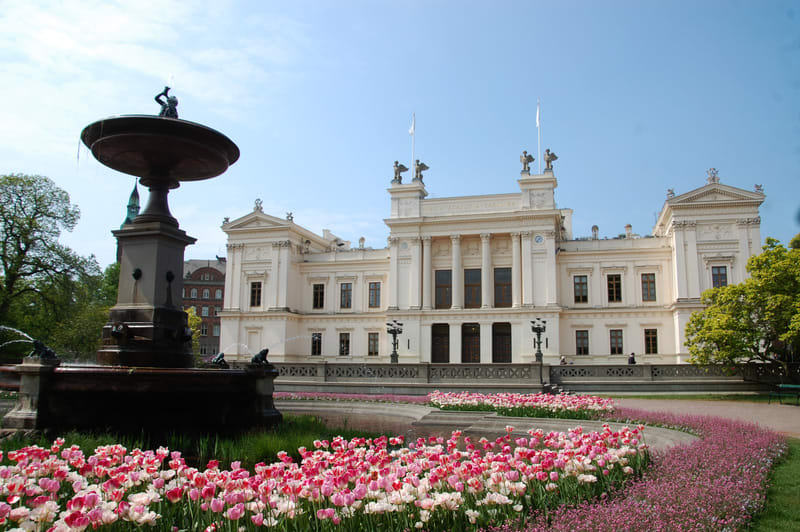 En bild som visar en stor, ljusbeige eller vit byggnad med klassicistisk arkitektur, inklusive kolonner, gesimser och statyer på taket. Framför byggnaden finns ett stort, mörkt bronsfärgat springvatten med flera nivåer, där vatten sprutar från flera
