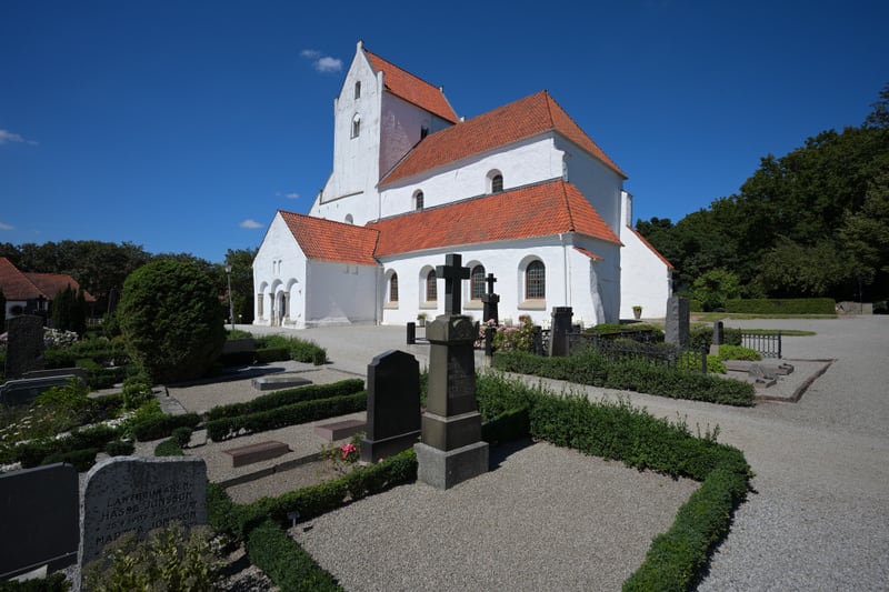 Bilden visar en vit kyrkobyggnad med orange tak i romansk stil. Framför kyrkan finns en kyrkogård med flertalet gravstenar, grusgångar och välklippta häckar. En del gravstenar är höga och monumentala, medan andra är lägre och rektangulära. En stor gr