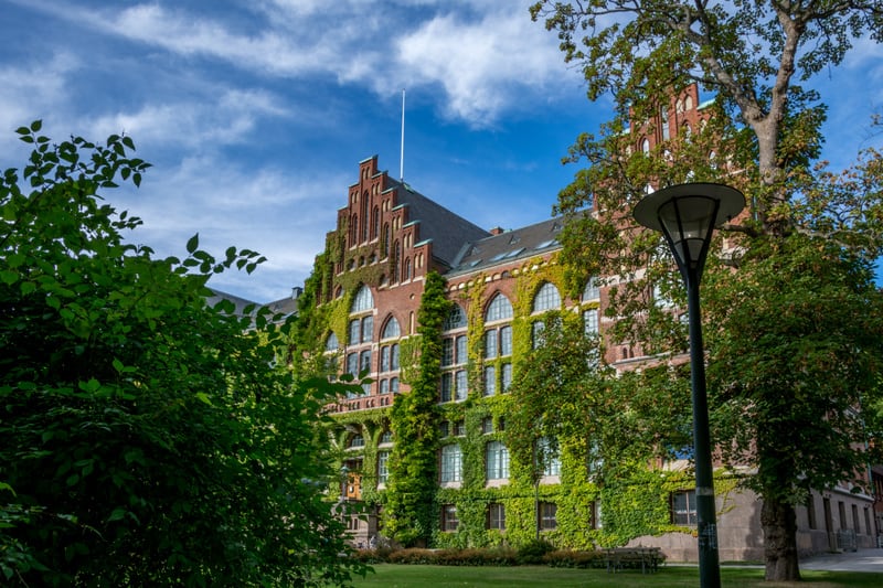 En stor, röd tegelbyggnad med många stora fönster dominerar bilden. Byggnaden är delvis täckt av grön murgröna. Den har ett brant tak med flera takfönster och gavelspetsar. Framför byggnaden finns en grön gräsmatta, och flera träd och buskar med grön