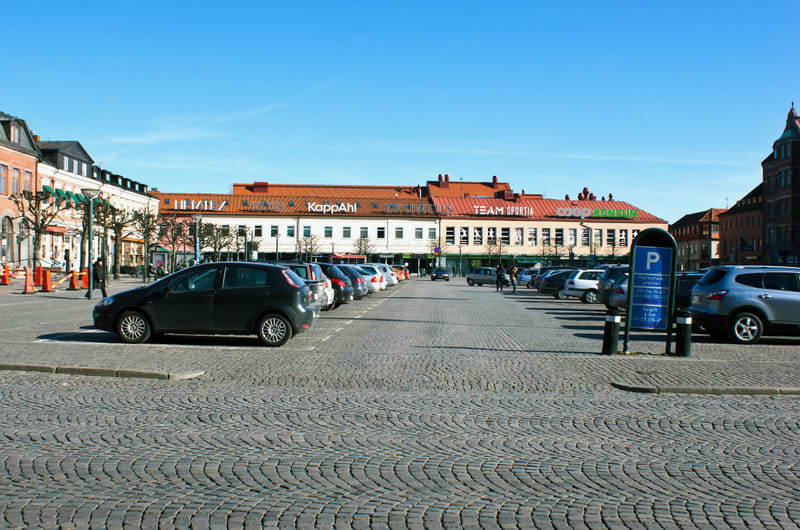 Bilden visar ett torg med kullerstenstäckning, där en stor parkeringsplats framträder i förgrunden och mitten. Ett flertal bilar är parkerade i rader på torget. I bakgrunden syns en stor byggnad med flera skyltar, bland annat "KappAhl", "TEAM SPORTIA