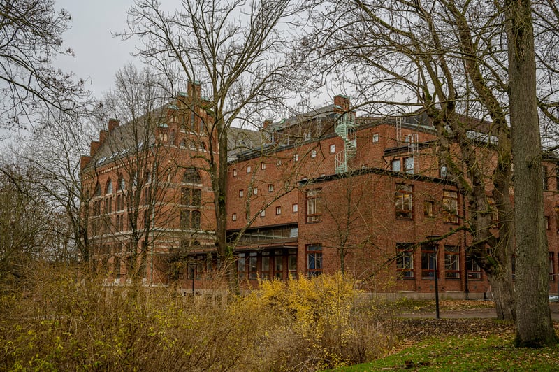 Bilden visar en stor byggnad i tegel sedd från ett grönområde. Byggnaden har flera våningsplan och är konstruerad med rött tegel. Den har många fönster av varierande storlek och form, samt tak i grönt eller gråaktigt material. Små, fyrkantiga fönster
