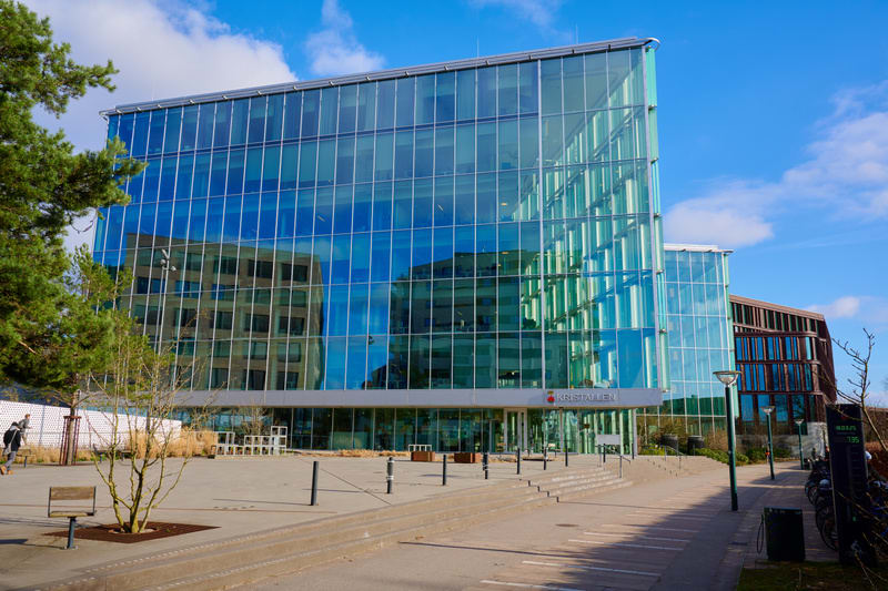 En modern byggnad med en fasad av glas dominerar bilden. Glasfasaden reflekterar delar av omgivningen, inklusive en annan byggnad och den blå himlen med vita moln. Framför byggnaden finns en öppen plats med grå markbeläggning, med några bänkar och pl
