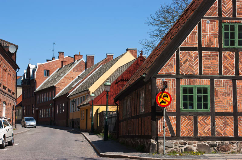 En gata i en stad med äldre byggnader. Till höger i bilden står en byggnad med fasad i tegel och mörka träbjälkar, samt fönster med grönmålad karm. Framför denna byggnad står en skylt med ett rött kryss över en bil och en motorcykel, vilket indikerar