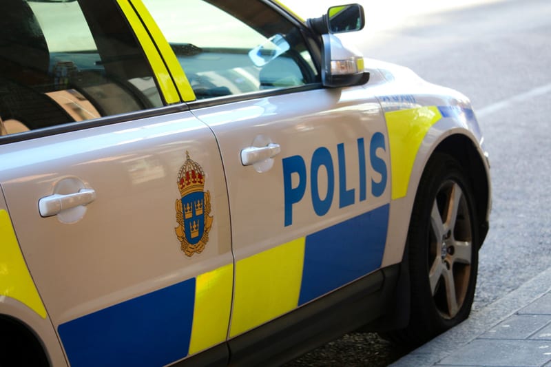 En beige polisbil är parkerad vid sidan av en trottoar. Större delen av bilens sida syns, med texten "POLIS" i blå versaler på dörren. Polisbilen har även gula och blåa ränder, samt en bild av lilla riksvapnet med tre kronor mot blå bakgrund. Baksätets fönster är svagt tonat och reflekterar omgivningen. En del av bilens framhjul är synligt.