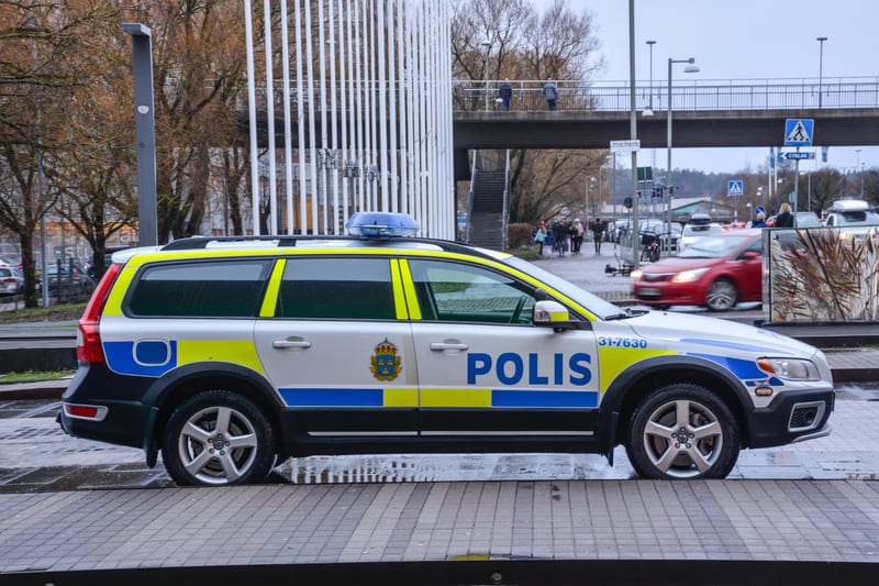 En svensk polisbil, en vit Volvo kombi med blå och gula reflexdetaljer samt texten 'POLIS' på sidan, står parkerad vid en trottoarkant. Bilen har en blå ljusramp på taket och förseglingsnummer '31-7630' på fronten. I bakgrunden syns en stadsmiljö med