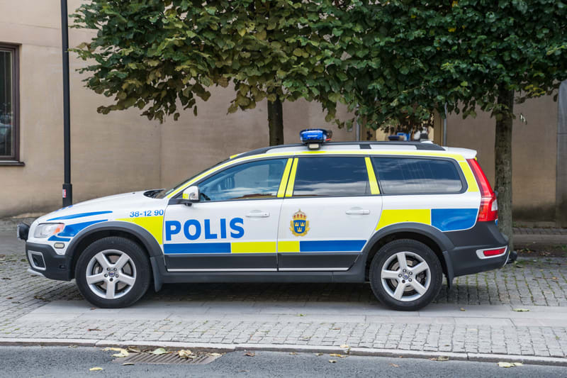 Bilden visar en svensk polisbil av modellen Volvo XC70. Bilen är vit med tydliga blå och gula reflexband längs sidorna, samt texten "POLIS" i blått på bilens sida. På framdörren finns även Sveriges lilla riksvapen. Bilen står parkerad på en trottoark