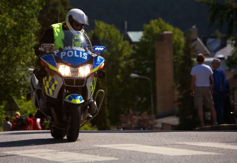 Bilden visar en polismotorcykel som rör sig framåt på en väg under dagtid. Föraren bär en vit hjälm och en gul reflexväst. Motorcykeln är blå, vit och gul med texten "POLIS" synlig på framsidan och har blåa och gula rutor samt fungerande strålkastare