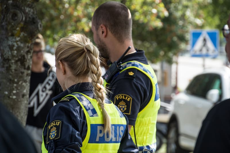 Bilden visar två polisuniformerade personer sedda bakifrån, troligen utomhus en dagtid. Den ena personen, med blont flätat hår, är i förgrunden och bär en blå poliströja med ett gult reflexväst över. På västen står "POLIS" skrivet med stora bokstäver