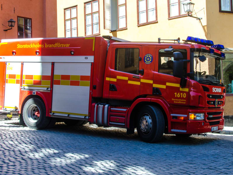 En röd brandbil modell Scania står parkerad på en kullerstensgata. Brandbilen har texten "Storstockholms brandförsvar" samt numret "1610" tryckt på sidan. Den har blåa utryckningsljus på taket och i fronten. På sidan av fordonet syns luckor och panel