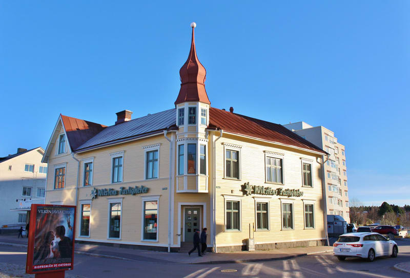 Bilden visar en gul tvåvåningsbyggnad med rött tak och en spetsig takkupol i ett hörn. Byggnadens fasader är klädda med liggande träpanel. På hörnet finns en ingång, och på övre våningen ovanför den ingången finns ett burspråk. Skyltar med texten "Wi