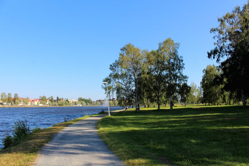 Bilden visar ett landskap med en bred vattenyta, sannolikt en sjö eller större flod, till vänster. På högra sidan av bilden syns en gång- eller cykelväg som följer vattenkanten. Längs gångvägen och i bakgrunden till höger finns en stor grön gräsyta m
