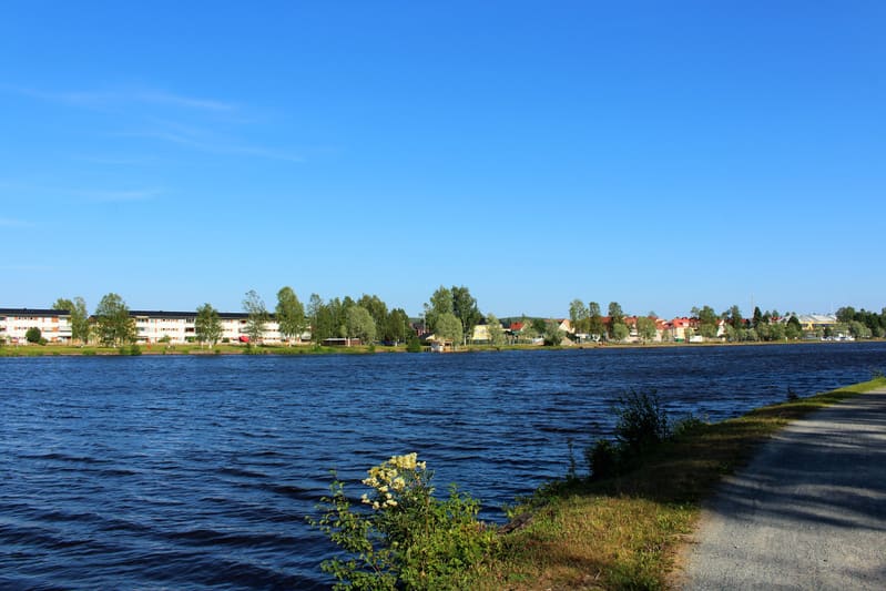 En vidvinkelbild tagen utomhus under dagtid med en stor del av en sjö eller större vattendrag i förgrunden och mitten. Vattnet är mörkblått med synliga vågor. På andra sidan vattnet syns en bebyggelse med både flerbostadshus och villor, omgivna av gr