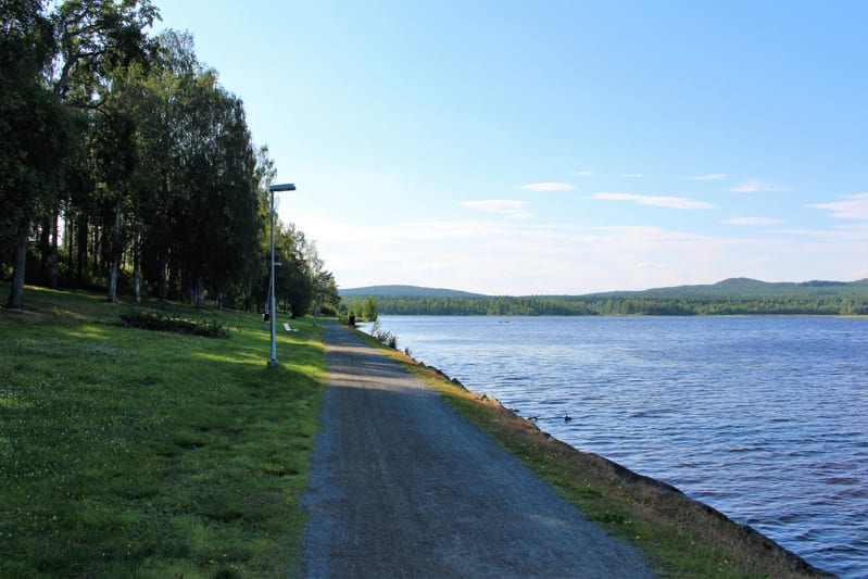 Bilden visar en grusväg längs ett vattendrag under dagtid. Till vänster om grusvägen finns en gräsmatta som övergår i en skog fylld med lövträd och barrträd. Längs grusvägen står flera lampstolpar och en vit bänk syns i bakgrunden. Vattendragets yter