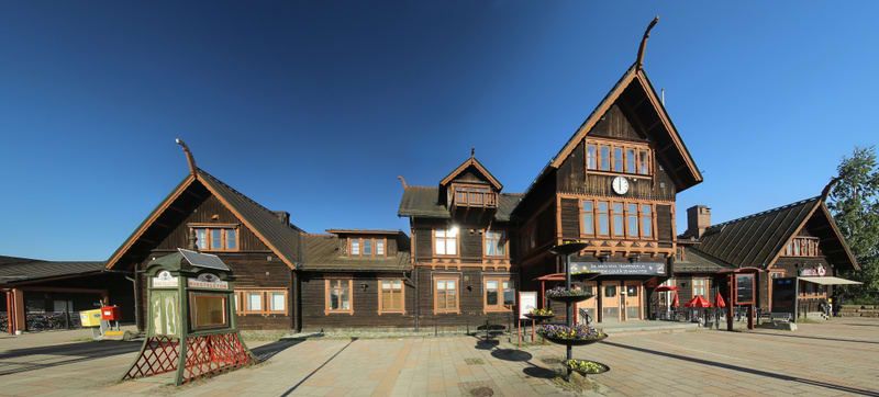 Bilden visar en panoramvy över en stor, mörkbrun träbyggnad med distinkt nordisk arkitektur under en klarblå himmel. Byggnaden har flera sektioner med olika takhöjder och spetsiga tak, varav några är dekorerade med snidade detaljer. På den högre dele