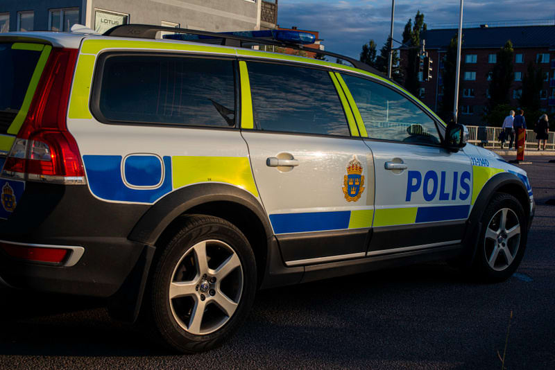 Bilden visar sidan av en svensk polisbil parkerad utomhus. Bilen är en vit Volvo kombi med blå och gul reflekterande dekor. På sidodörren syns texten "POLIS" i blått, samt svenska riksvapnet. Takräcken och blå ljusramper är synliga på taket. I bakgru