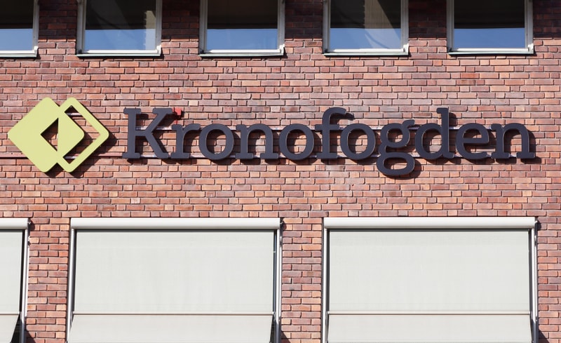 Bilden visar fasaden på en tegelbyggnad med texten "Kronofogden" i mörk färg och en grön symbol med två sammanlänkade rombformade figurer. Ovanför texten syns två rektangulära fönster med vita karmar som reflekterar ljus. Under texten finns minst två fönster med neddragna, ljusgrå persienner eller rullgardiner som täcker hela fönsterytan. Tegelfasaden är i en rödbrun nyans och belyses av solljus som skapar skuggor bakom bokstäverna och symbolen, vilket indikerar att bilden är tagen utomhus under dagtid.