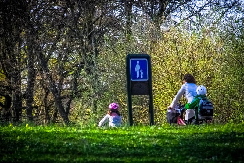Bilden visar en person på cykel som färdas bort från betraktaren, med ett barn i en cykelsits på pakethållaren. Till vänster om cykeln sitter ett annat barn, som bär hjälm, i en gräsbacke. Mellan barnet i gräset och cykeln står en skylt med en symbol