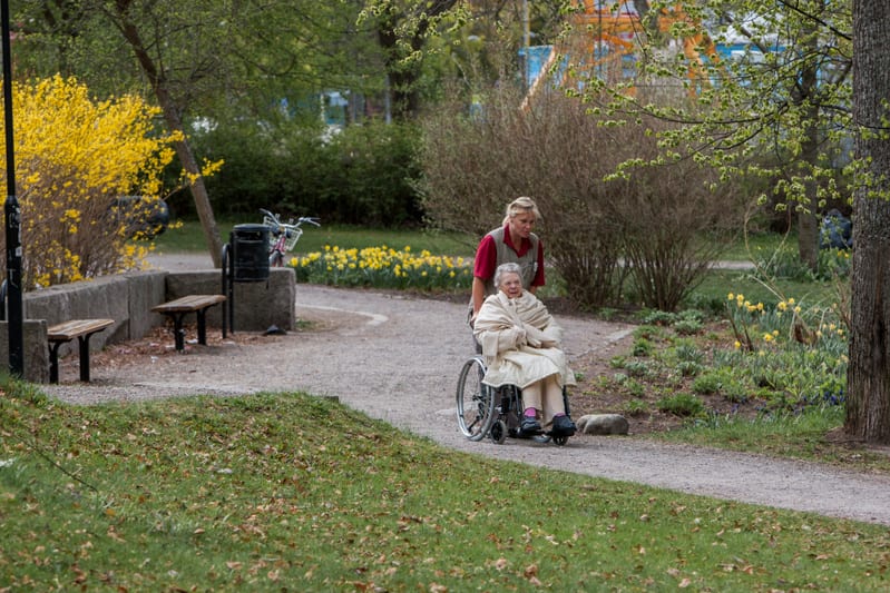 Bilden visar en kvinna som skjutsar en äldre kvinna i rullstol på en grusgång utomhus. Båda personerna är vända bort från kameran men den äldre kvinnan i rullstolen tittar mot vänster. Kvinnan som skjutsar är klädd i en röd kortärmad tröja. Den äldre