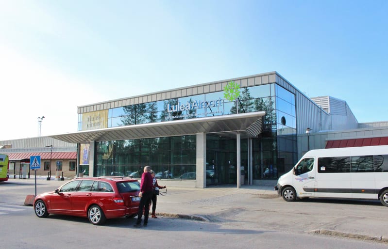 Bilden visar entréområdet till Luleå Airport en solig dag utomhus. I centrum av bilden står en röd kombibil parkerad, med två personer som står vid bagageutrymmet. Till höger om bilen ses en vit minibuss. I bakgrunden syns flygplatsbyggnaden med stor